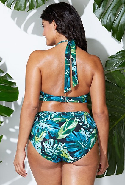 Woman wearing a tropical leaf print bikini against a white wall with green leaves.