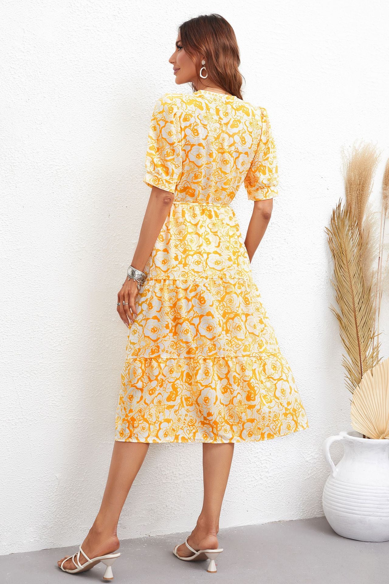 Woman wearing a yellow floral dress standing against a white wall with a decorative plant.