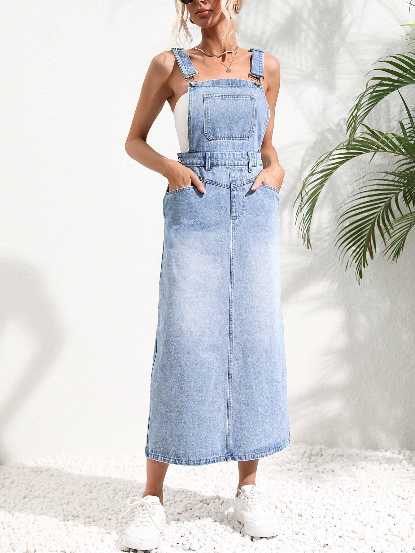 Woman wearing a light blue denim dress against a white background with a plant on the right. Women wearing tight waist strap mid-length denim dress with kurti