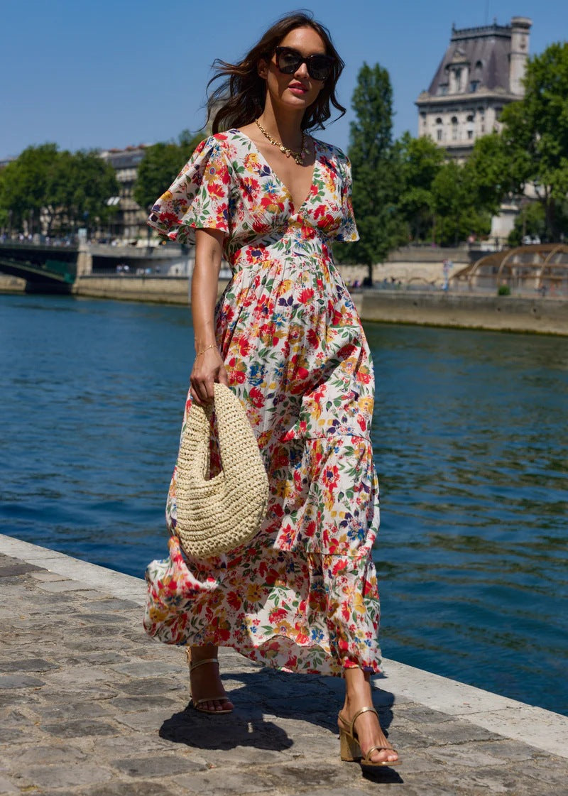 Woman in a colorful floral dress standing by a waterfront with trees and buildings in the background.