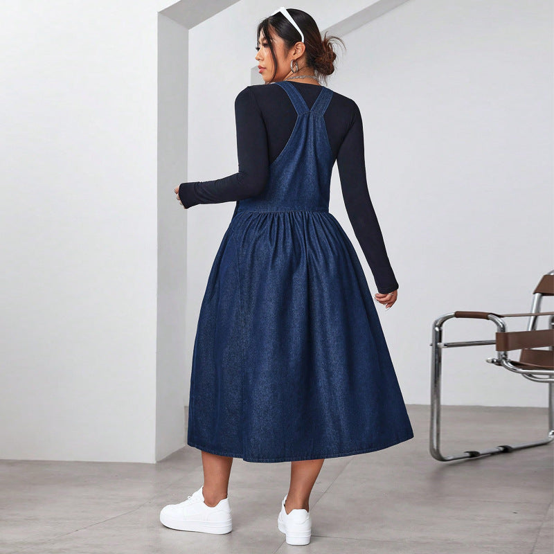 Woman wearing a blue denim pinafore dress with a black long-sleeve shirt underneath, standing in a minimalistic room.
