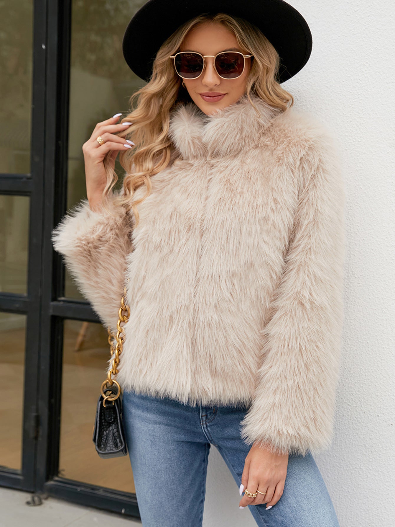 Woman wearing a beige fluffy jacket, black hat, and sunglasses outdoors.